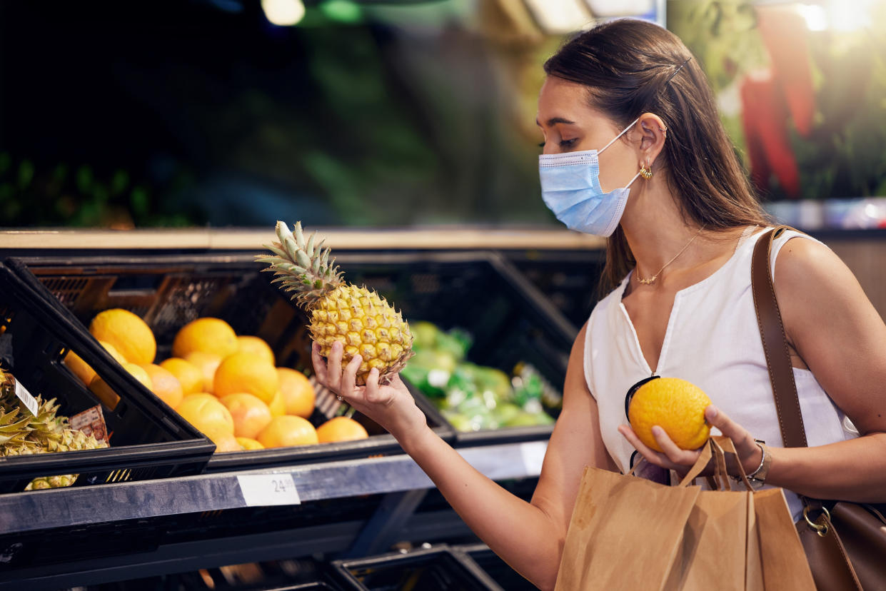 A woman wearing a medical mask in a grocery store examines a pineapple.