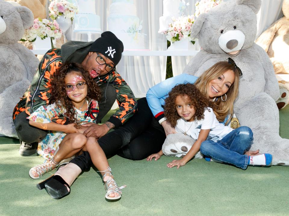 nick cannon holding daughter monroe cannon, and mariah carey holding son moroccan cannon, sitting on a green floor and smiling with large plush bears in the background