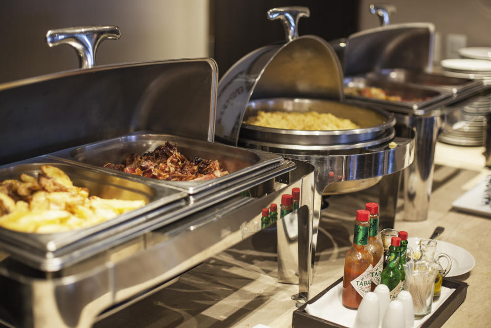 A buffet setup featuring trays of various foods, including roasted potatoes, bacon, and pasta. Hot sauce and condiments are also visible at the front