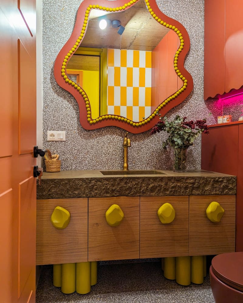 Bathroom with wavy orange and yellow mirror, wood cabinets with stone slab counter and yellow irregular shaped knobs