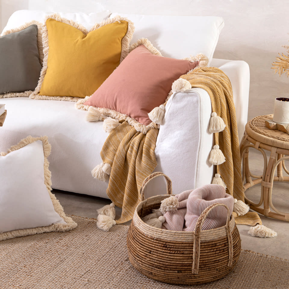 Muse Chester Feather Cushions with fringes in pink, ochre and grey sit on a cream couch with wicker table and basket.