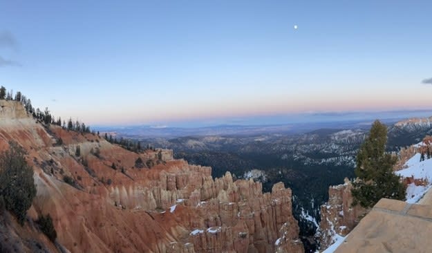 A tiny moon on top of the skyline looks down onto the trees-lined, sparse rock formations of Bryce Canyon.