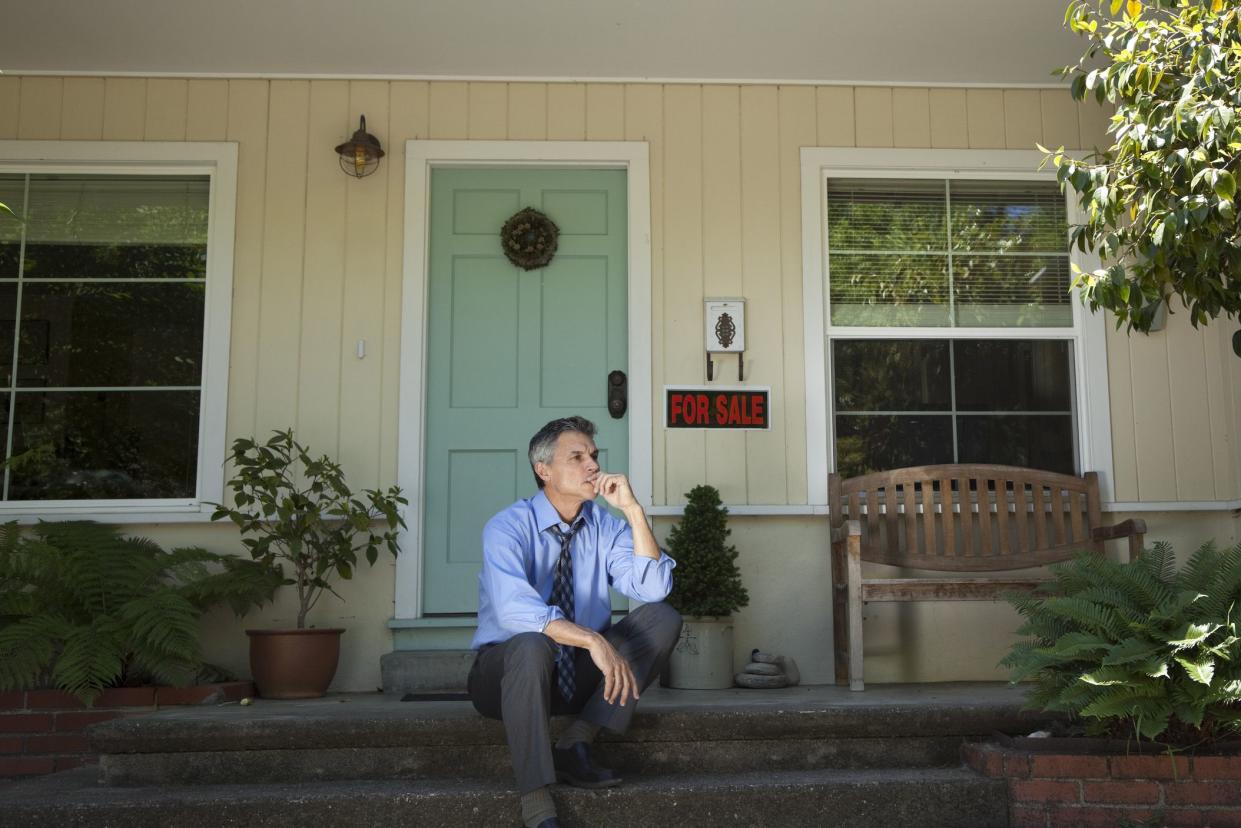 mature man in suit  with sad expression sitting outside house with for sale sign