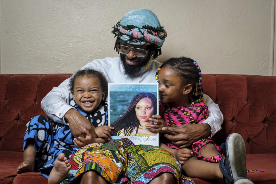 In this photo provided by Henry Danner, Omari Maynard sits with his children, Khari, left, and, Anari, holding a photo of their late mother, Shamony Gibson, at home in the Brooklyn borough of New York on April 9, 2022. Gibson passed away in 2019, two weeks after giving birth to Khari due to a pulmonary embolism. “She wasn’t being heard at all,” said Maynard, an artist who now does speaking engagements as a maternal health advocate. (Henry Danner via AP)