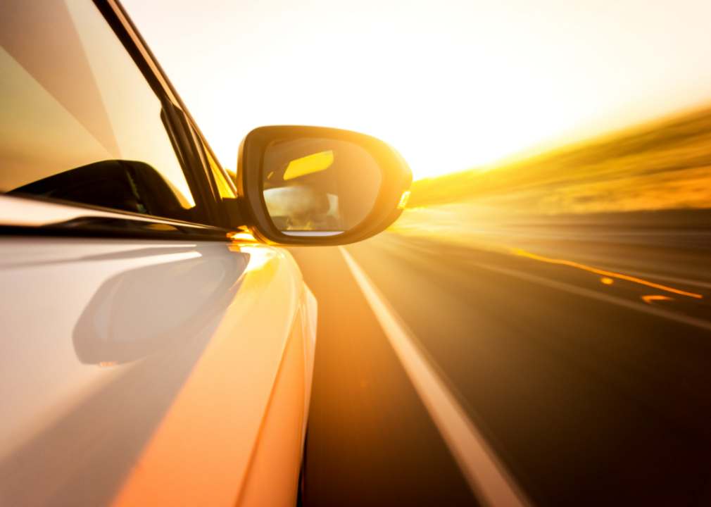 The side mirror of a white car speeding down the highway at sunset.