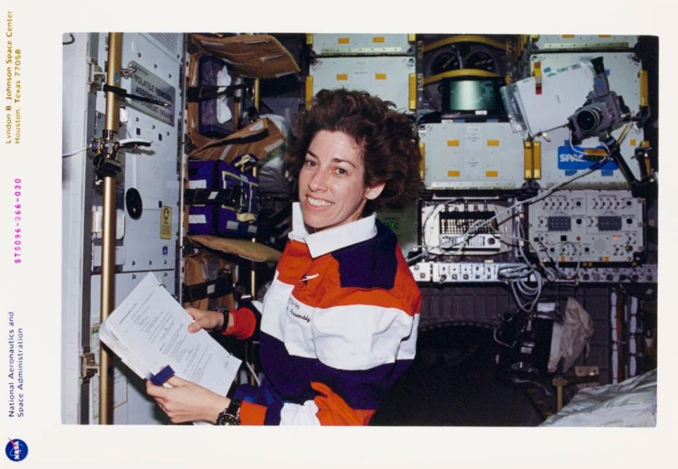 ellen ochoa smiles at the camera while floating in a space shuttle, she wears an orange, blue, and white striped polo shirt and watch and holds a piece of paper in both hands, around her is lots of technical equipment