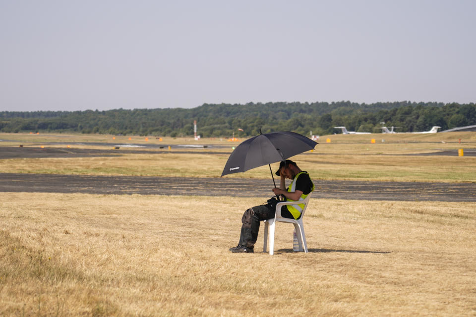 A member of the staff shelters from the sun under an umbrella as he sits next to the runway, at the Farnborough Air Show fair in Farnborough, England, Tuesday, July 19, 2022. Some 1191 exhibitors from around the world show their newest developments in Future Flight, Space, Defence, Innovation, Sustainability and Workforce from July 18th until July 22, 2022. (AP Photo/Alberto Pezzali)