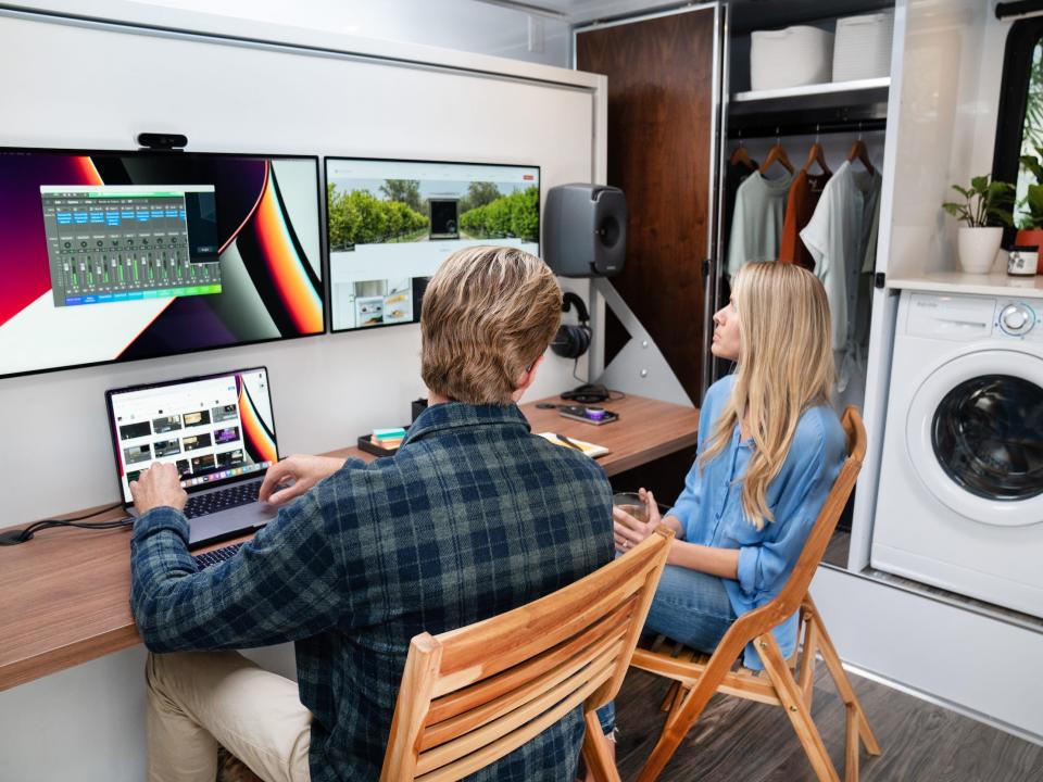 Two people sitting at a desk with Apple equipment. There's a washer and closet nearby.