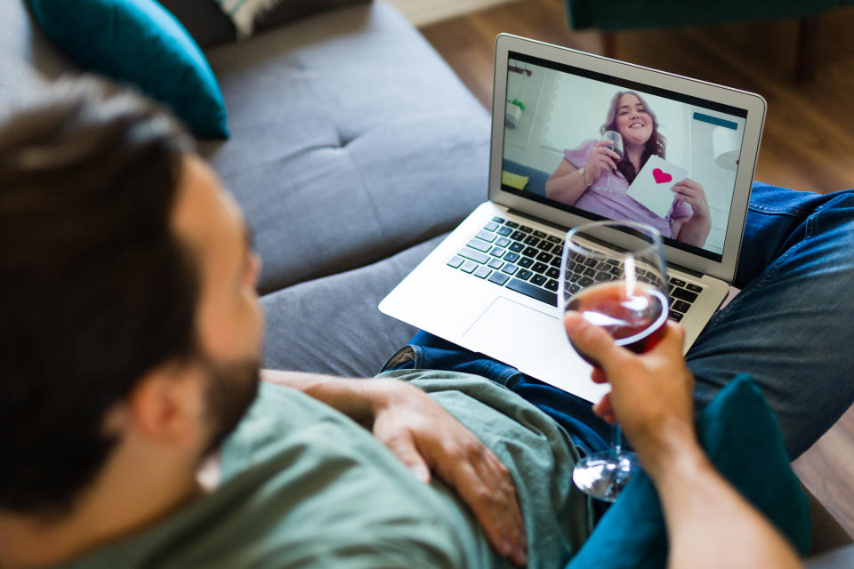 A man sits on his sofa with a glass of wine in one hand while looking at his laptop, where he is on a video call with his girlfriend, who is on the screen holding a glass of wine and a card with a heart