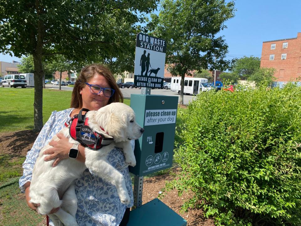 Taunton PD's comfort dog, Maggie, being held by her handler, Katrina Lee, Co-Response Clinician for the Police Department. They are standing next to one of 4 dog waste stations installed around the downtown area. This dog waste station is located in the Mill River park/walking path area, which is adjacent to Spring St. and the parking lot behind the police station. Photo taken July 26, 2023.