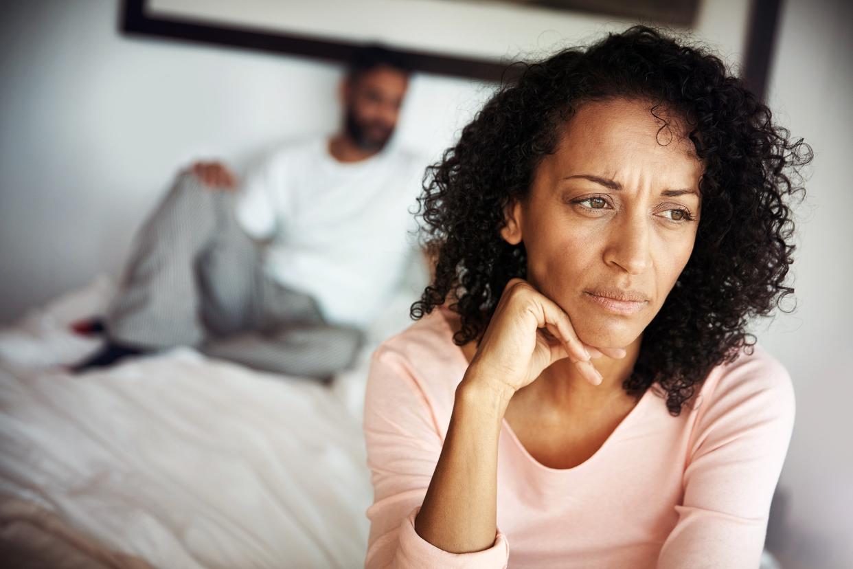 Depressed woman sitting up in bed with her husband in the background