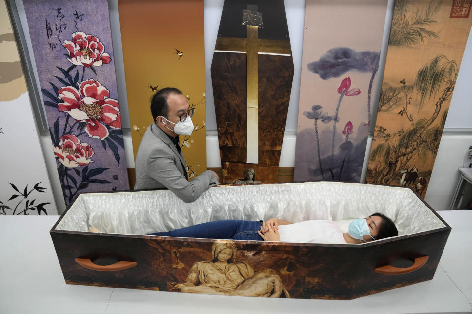 Wilson Tong, CEO of LifeArt Asia, left, talks to a reporter who tries out a catholic-designed paper coffin at Tong's factory in Hong Kong, Friday, March 18, 2022. Hong Kong is running short of coffins during its deadliest outbreak of the coronavirus pandemic, which has cost about 6,000 lives so far this year. LifeArt, a company in Hong Kong is trying to make an alternative, cardboard coffin, which it says is environmentally-friendly. (AP Photo/Kin Cheung)