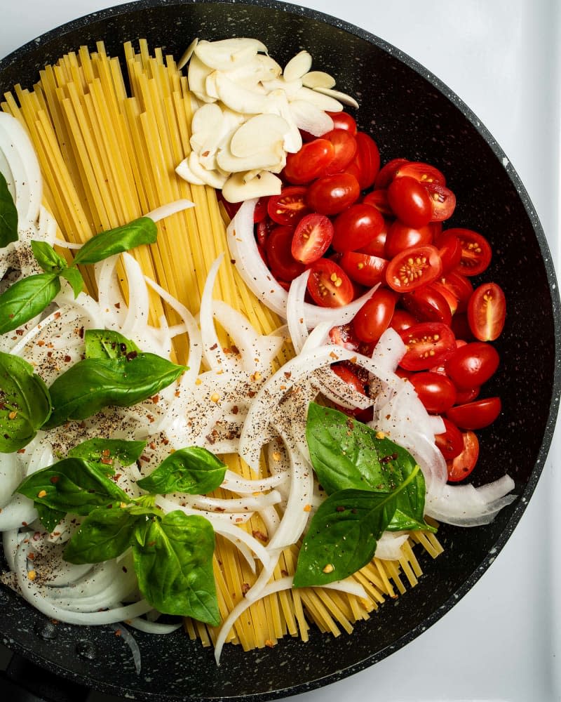 basil, cherry tomatoes, dried pasta, garlic cloves, and white onion seasoned and ready to cook in a cast iron skillet