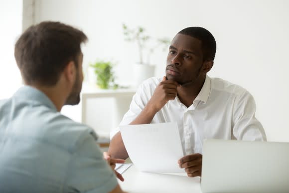 Man sitting across from another man, holding a piece of paper and looking thoughtful.