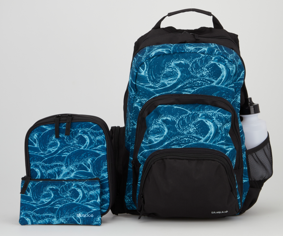 A dark blue and light blue backpack with a wave design stands on a white background on the right with the added smaller lunchbox and pencil case in the same fabric stand on the left. 