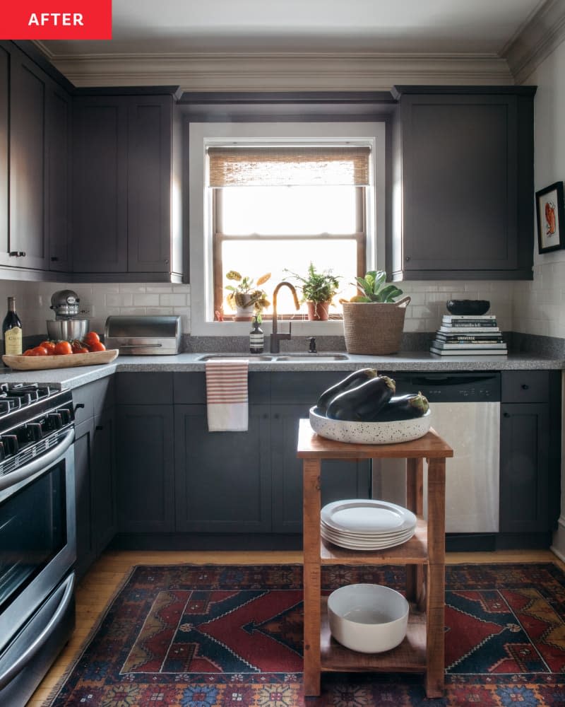 sheer roll down curtain, dark grey blue cabinets, dishwasher, sink, window, plants, red and blue patterned rug, small wood 2 shelf island, white bowls, oven, red and white dish towel, tomatoes on cutting board