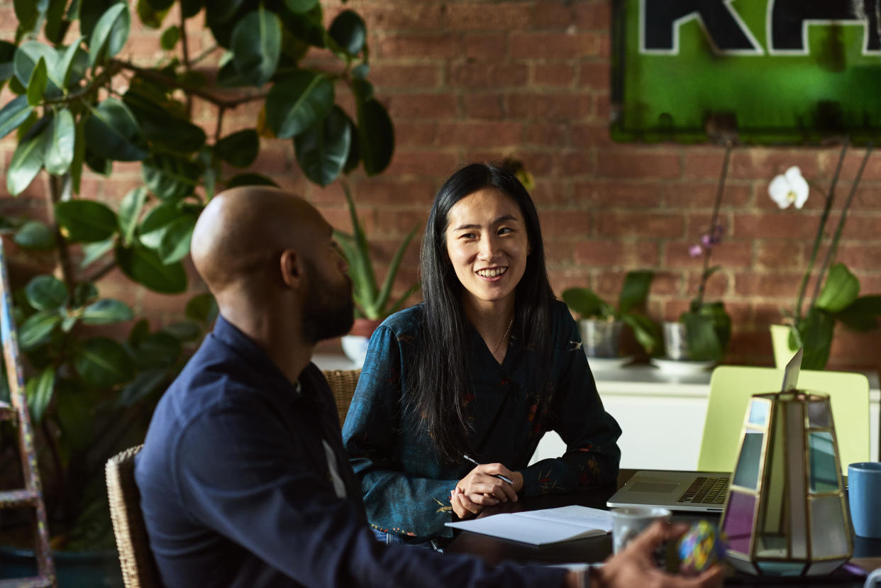 Two creative designers sitting in meeting in modern office, woman with note paper and pen listening to man with cheerful expression