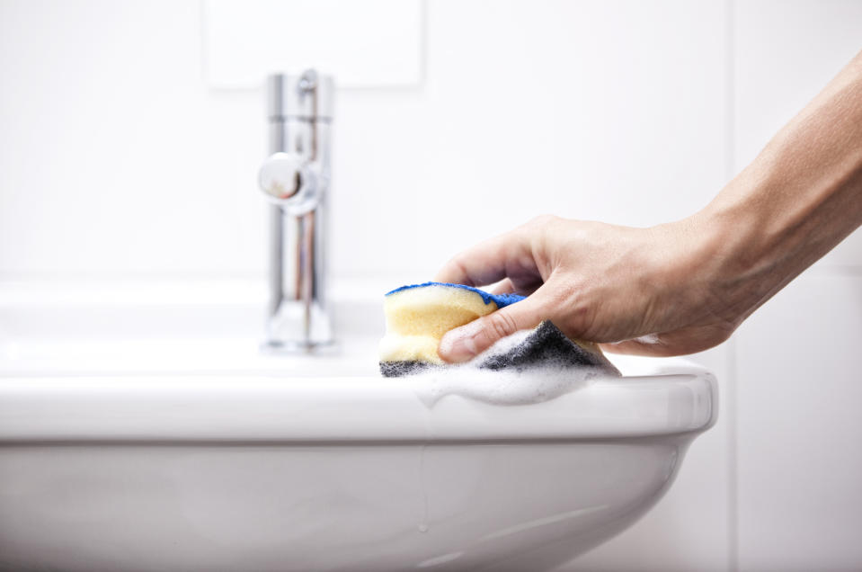 A hand is cleaning a white sink with a sponge, producing some soap suds