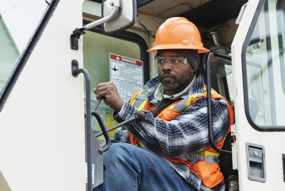 Man operating heavy machinery, wearing a plaid shirt, safety vest, and orange helmet on a construction site