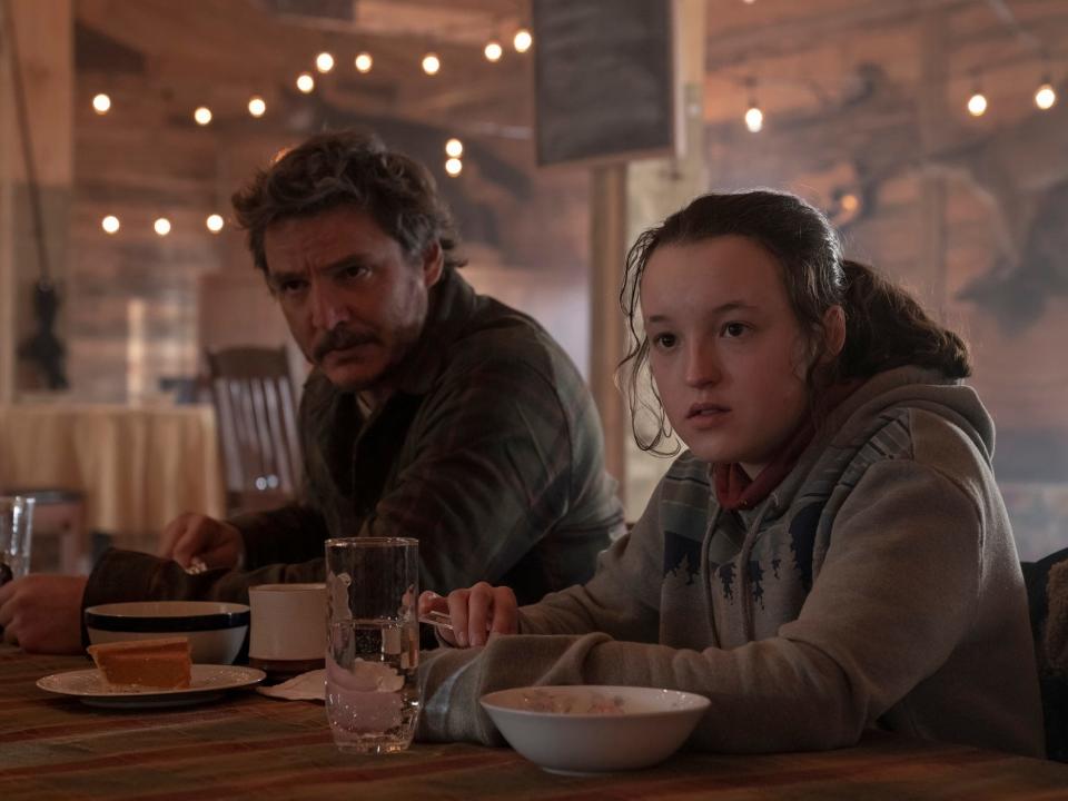 joel and ellie in the last of us. they're sitting at a dining table inside a large wooden cabin lodge, eating soup, drinking water, and looking very seriously to the side