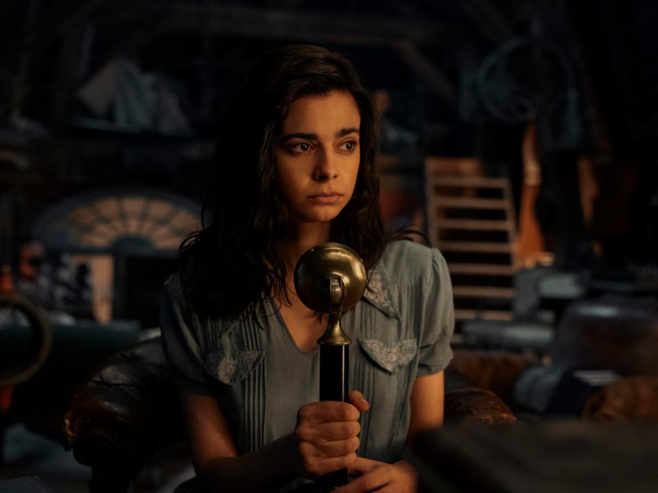 aria mia loberti as marie-laure in all the light we cannot see. she's wearing a blue dress, and holding a radio. microphone, looking concerned with her face half lit