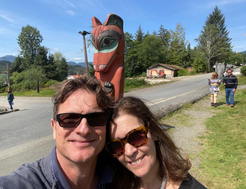 A woman and man take a selfie in front of a totem pole.