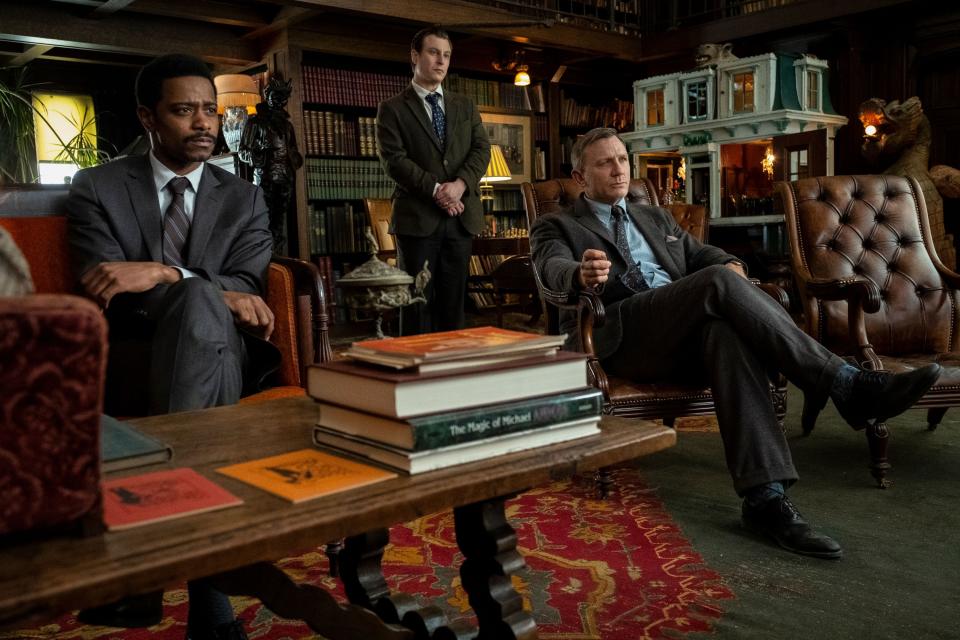LaKeith Stanfield, Noah Segan, and Daniel Craig sit in a library, engaged in conversation. LaKeith and Daniel sit in leather chairs, with books and decor around