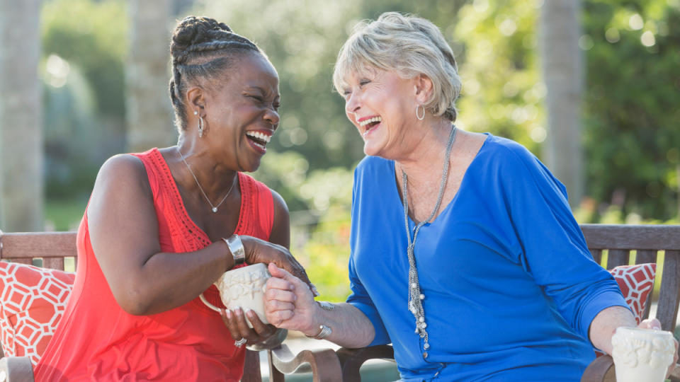 Two women sitting on a bench holding mugs while laughing together