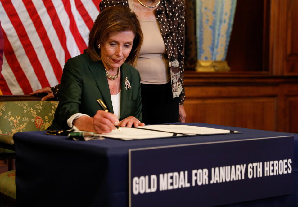 Nancy Pelosi holds a pen while seated at a signing table with a banner reading 