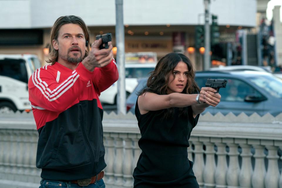nick zano as chad mcknight and shelley hennig as ava winters in obliterated, both intently aiming their guns while standing near a road