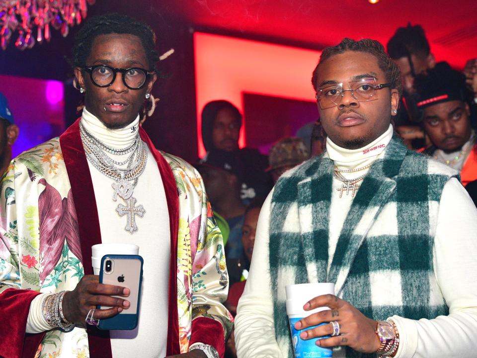 Young Thung, in a white turtleneck, glasses, and a silk robe, poses for a photo with Gunna, in a white turtleneck, glasses, and plaid vest.