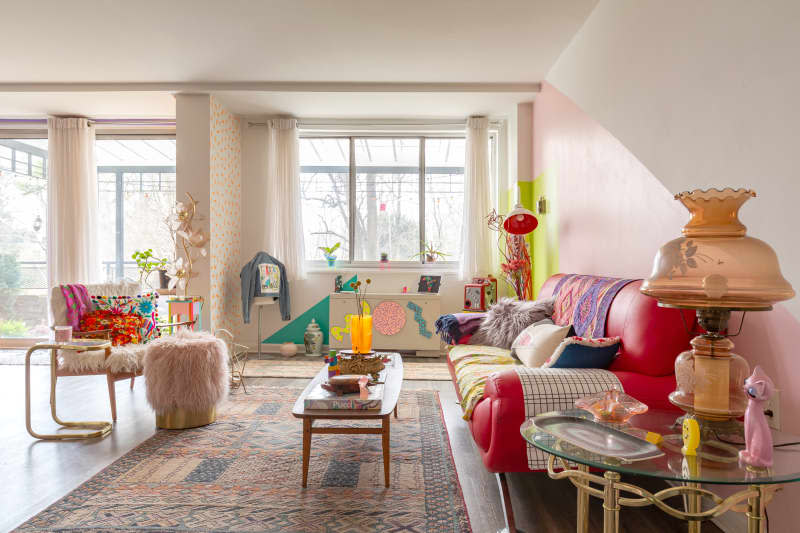 Colorful living room with red sofa, patterned throw pillows, large windows, pink, white, chartreuse, and lavender walls,  wood accent chair with shag throw, wood coffee table with books and colorful design objets