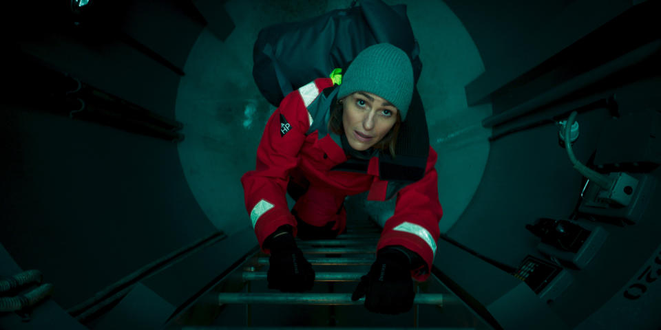 A woman in a red jacket and grey hat climbs up submarine ladders.