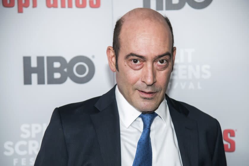 A man in a black blazer, white dress shirt and blue tie looks into the camera