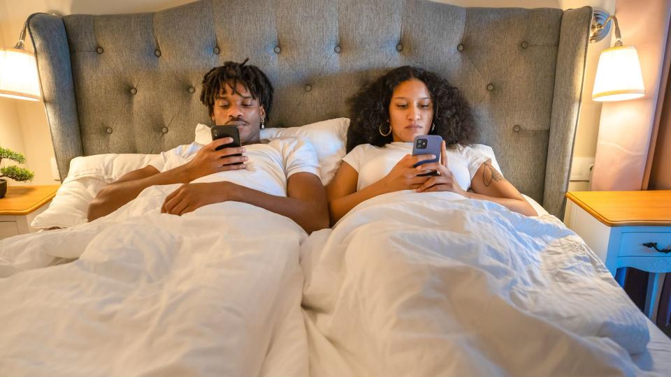 man and woman lie in bed next to each other, not speaking and both on their phones