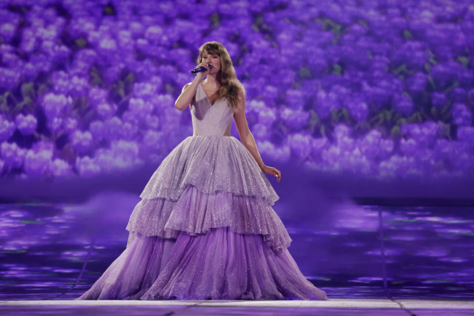 Taylor Swift performs on stage in a layered gown with floral background