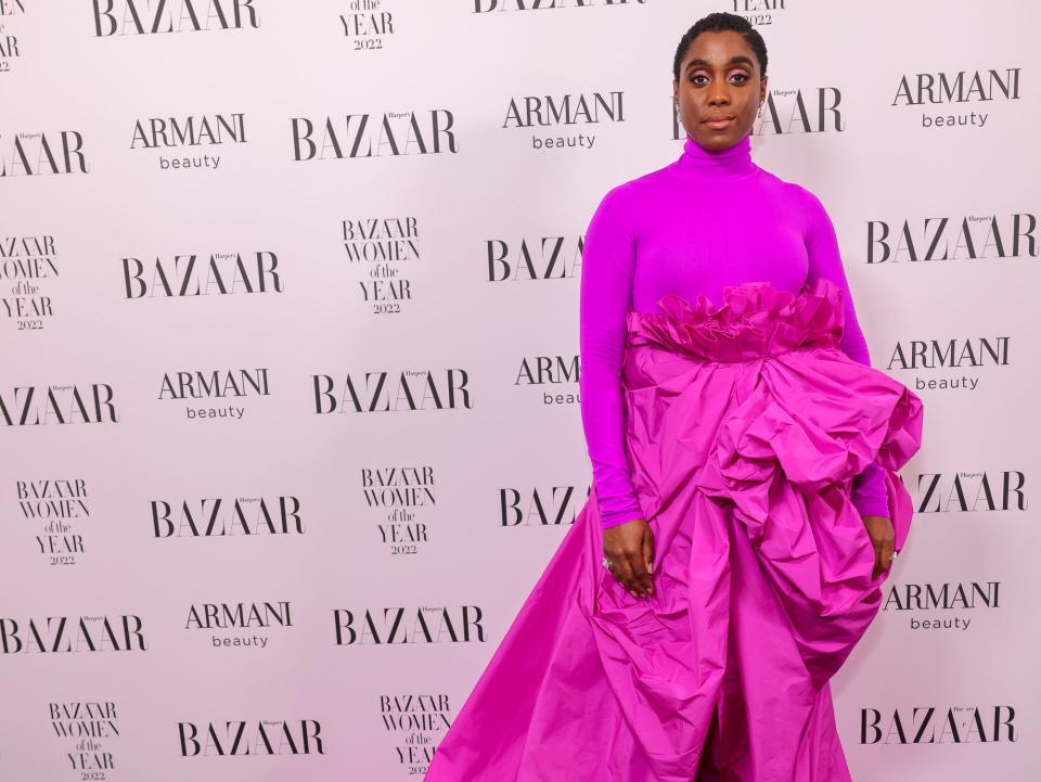 Lashana Lynch in pink/purple turtleneck and skirt