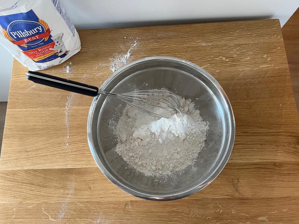 A silver bowl with flour, baking soda, and baking powder on a wood surface.