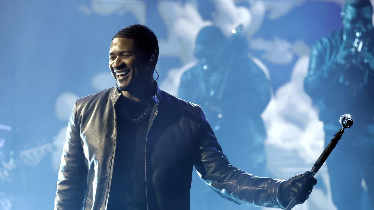 usher smiles while standing on a stand and holding a tilted microphone stand with one hand, he wears all black including a matching leather jacket and pants with a black t shirt
