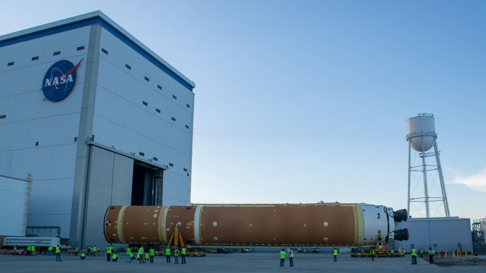 Eine große orangefarbene Raketenverstärkerstufe steht horizontal auf Rädern vor einem großen Hangar und Wasserturm.