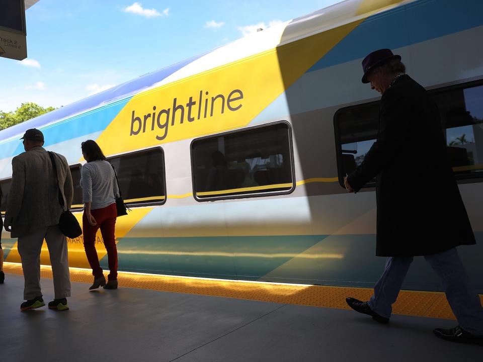 A multicolored Brightline train sits on a track with several passengers walking on the platform in front of it.