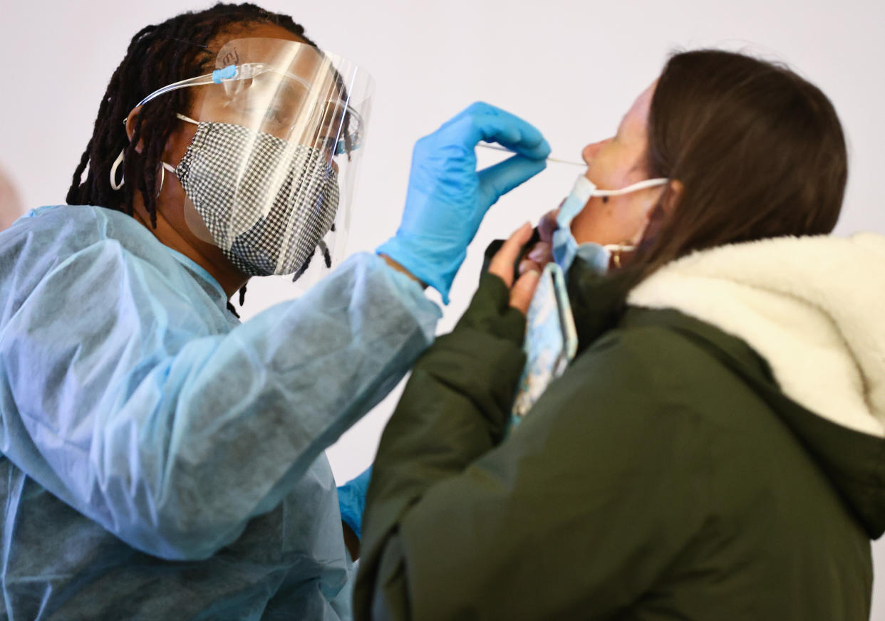 A woman in protective gear, including a mask, face shield, gown and gloves, administers a COVID-19 nasopharyngeal swab to another woman at a testing site.