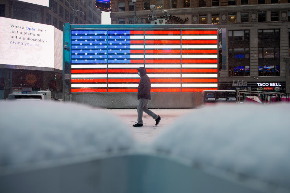 NEW YORK, NEW YORK - FEBRUARY 18: A person wearing a mask walks past a large American flag near piles of snow during a snow storm in Times Square on February 18, 2021 in New York City. The U.S. National Weather Service issued a winter weather advisory that a total of 6 to 8 inches of snow is expected on Thursday and Friday in parts of the Northeast, including Southeast New York. (Photo by Alexi Rosenfeld/Getty Images)