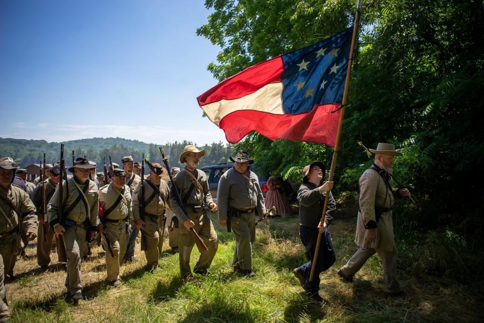 A crowd of Civil War reenactors walk with the official flag of the Confederacy.