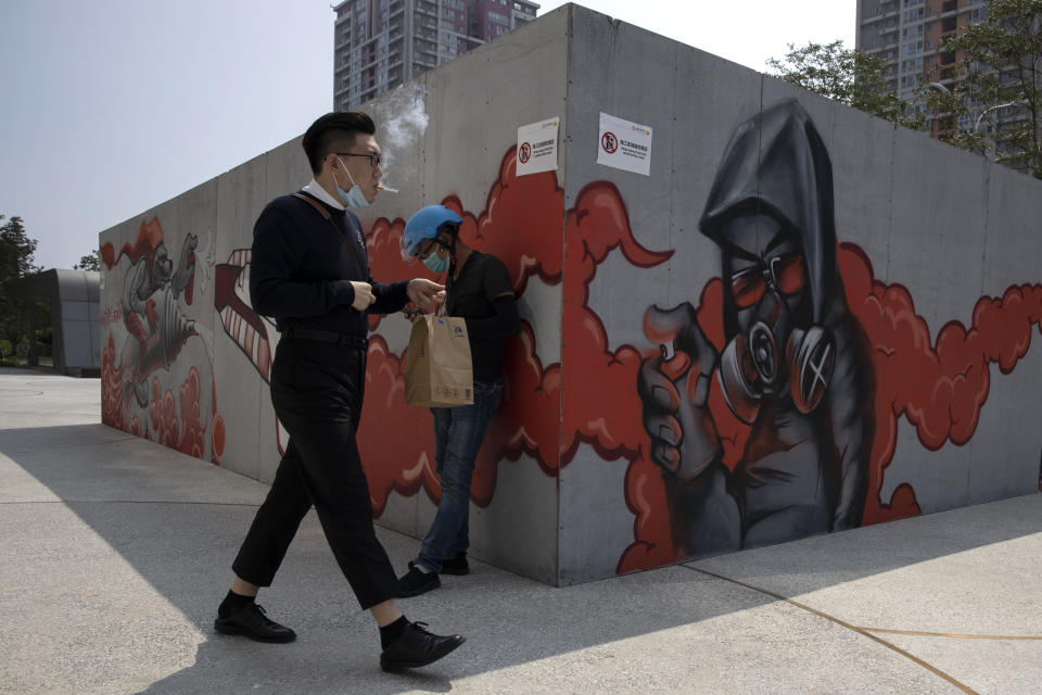 A man lowers his mask to smoke as he walks past graffiti artwork in Beijing Monday, July 13, 2020. China reported eight new cases, all of them brought from outside the country, as domestic community infections fall to near zero (AP Photo/Ng Han Guan)