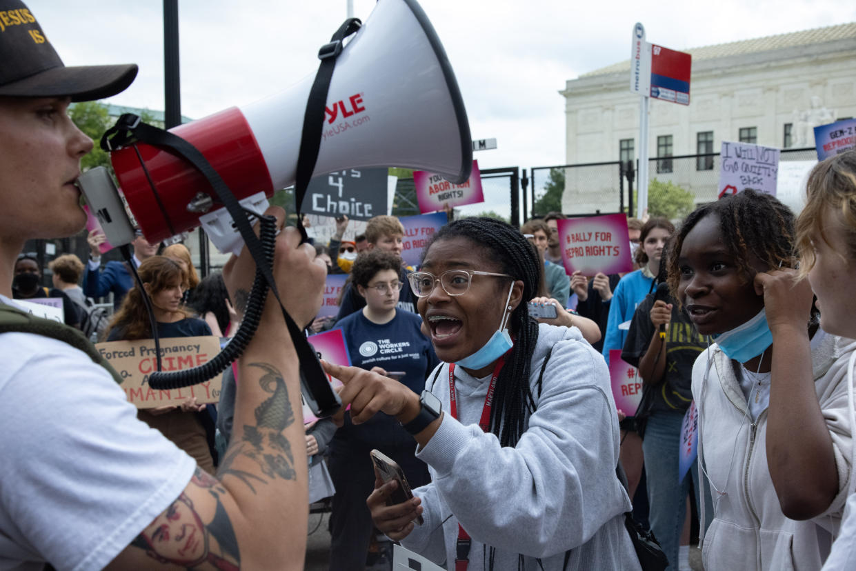 Two young Black women with blue surgical masks pulled down below their chins yell at an unmasked white man with tattoos speaking into a bullhorn. 