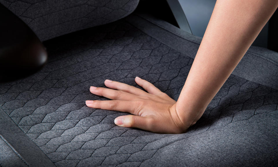 Marketing image for a product showing a person's palm pressing down on a gray patterned ergonomic chair.  Close-up of chair seat.