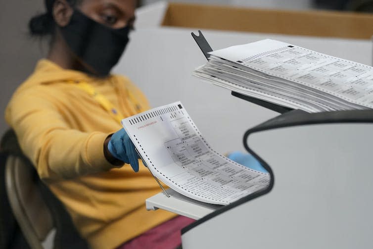 A person sits by a machine with a stack of ballots.