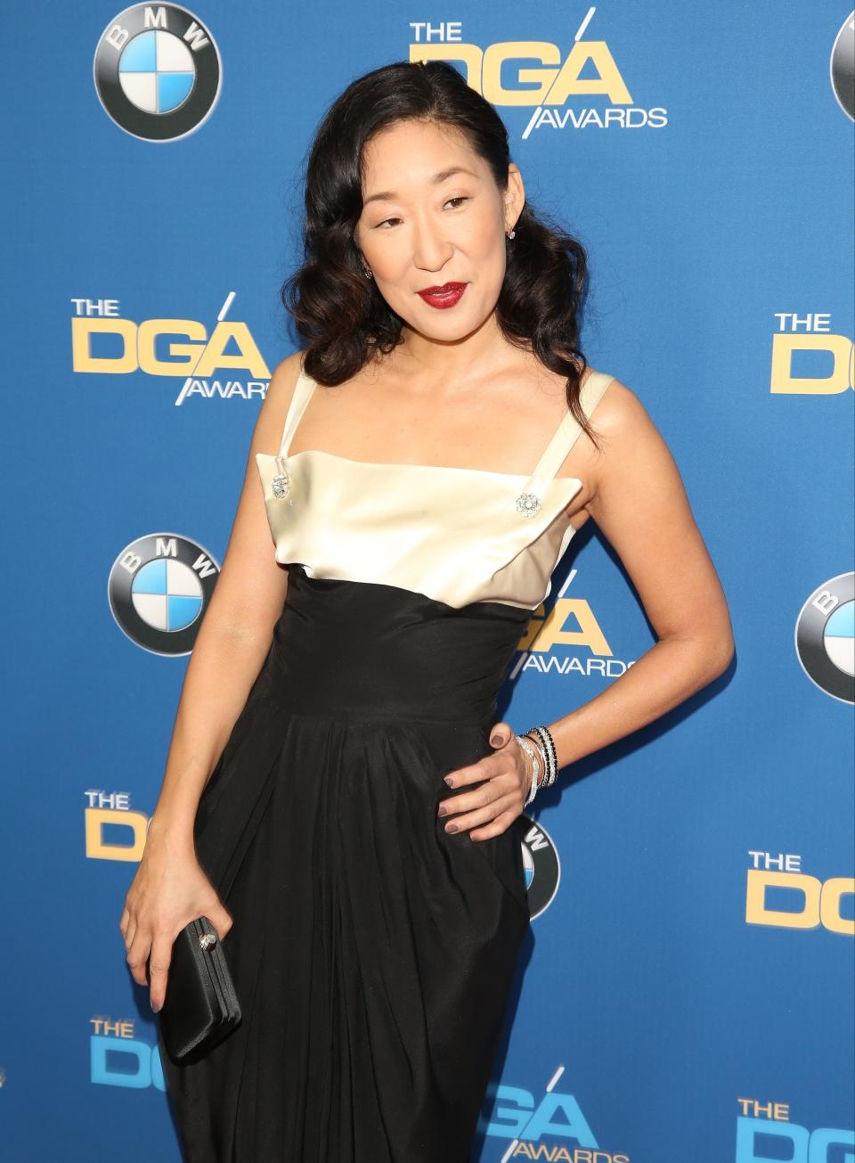 Sandra Oh on the red carpet at The DGA Awards wearing a sleeveless gown with a structured top and a flowing skirt, holding a black clutch