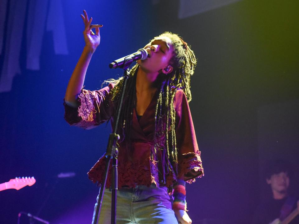 WILLOW on stage in a maroon wrap top with floral lace detailing, and blue jeans, singing into a mic. Her hair is in locs with a half up half down do.